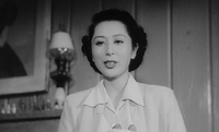 Una atractiva joven japonesa, vestida con una blusa blanca, aparece hablando, fotografiada desde abajo; al fondo a la izquierda se aprecia una lámpara, unas botellas sobre una repisa de la chimenea y parte de un cuadro.