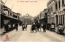 Photo ancienne montrant une rue dans laquelle circulent des pousse-pousses, avec en arrière-plan un grand bâtiment.