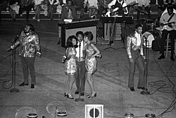 Three dark-skinned men and two dark-skinned women singing on a stage. Various musicians are visible behind them.