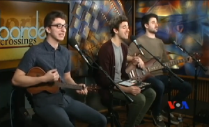 The members of AJR performing. From left to right, Ryan holding a ukulele, Jack singing, and Adam holding a guitar.