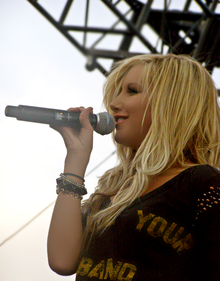 Tisdale, on a stage, sings into a microphone, holding it with her right hand. She is wearing a black blouse with the words "Your" and "Band" printed on it in yellow and few bracelets on her right arm.