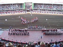 Approximately 50 people in red, black and white uniforms stand on a stage as a team of riders on horseback carry Canadian Flags in the background.
