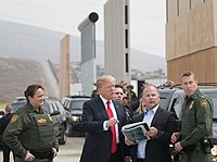 Trump speaks with U.S. Border Patrol agents. Behind him are black SUVs, four short border wall prototype designs, and the current border wall in the background