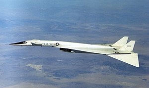 White delta-wing aircraft overflying mountains. The front of the fuselage features canard wings, and the wing tips are dropped.