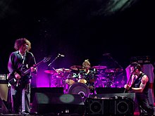 On a purple-lit stage, two men play guitars while a third man sits behind a drum kit.