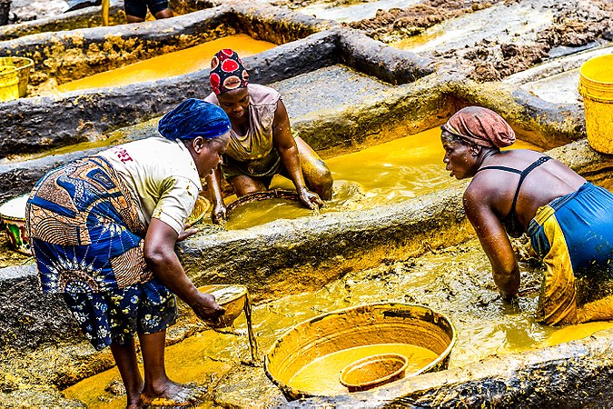 Photograph of three women in colorful garb manually refining palm oil within large rectangular indentions in the ground