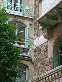 Photo en couleur d'une portion d'immeuble en angle rentrant avec encorbellement et parties métalliques : persiennes, hippocampe en enseigne et balcon avec masques en fer forgé