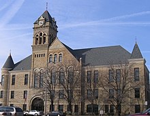 A large 3-story stone building built in 1895. 3-stories of windows line the front of the building with the two front corners containing cone shaped roofs that stick out from the main roof. Above the entrance is a large clock tower that is taller than the rest of the building.