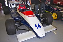 Photographie d'une monoplace de Formule 1 blanche, rouge et bleue, vue de trois-quarts, dans un stand d'exposition.