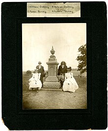 A group of four people split on either side of the grave of Ma-con-a-quah, also known as Frances Slocum. The people are labeled as William Godfroy, Mabel Bundy, Gabriel Godfroy and Victoria Bundy. Taken during the Slocum family reunion May 17, 1900.