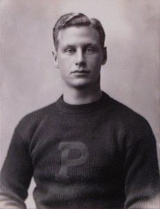 Черно-белое фото верхней половины молодого человека в свитере с большой буквой «П» спереди.