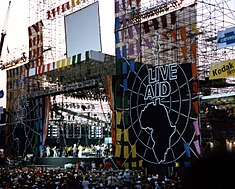 An outdoor stage with the a banner bearing the words "Live Aid" above an image of Africa and a large audience many raising their hands
