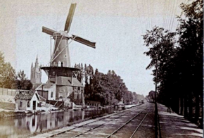 Photographie en noir et blanc d'une voie d'eau longée par une voie ferroviaire sur la rive droite, des habitations et un moulin ponctuant la rive gauche.