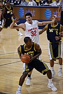 Баскетболист в темно-синей форме маневрирует с баскетбольным мячом, а защитник в белом находится в воздухе за его спиной.