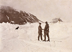 Two men shake hands in the midst of a snowfield, with a dog sitting nearby. Dark hills are shown in the background.