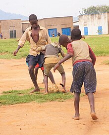 Fünf Jungen spielen barfuß auf sandigem Boden Fußball. Ihre Blicke sind nach unten auf den Ball gerichtet. Einer der Jungen ist deutlich größer als die anderen. Hinter ihnen befindet sich eine Rasenfläche und dahinter fensterlose Gebäude mit Flachdächern.