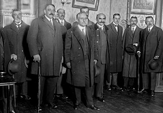Photo en noir et blanc d’un groupe d'hommes d'un certain âge, moustachus ou barbus, chapeaux à la main, portant de longues vestes foncées et des cravates