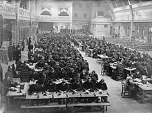 Monochrome photograph showing a hall full of recruits sat at tables. They each sit in front of Morse code receiving equipment and shown wearing headphones on their ears. There are a number of instructors shown standing to the left of the picture. All the recruits are dressed in air cadet uniform.