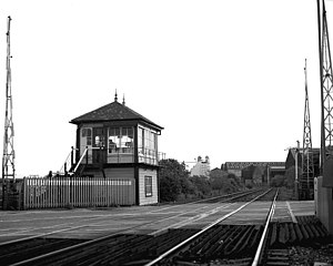 Raised cabin on left of railway lines stretching into distance on a left curve with large commercial buildings behind