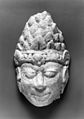 Among the stuccos recovered from various ancient sites in Nakhon Pathom, there is a type with tiered conical headdress consisting of many triangular elements. This head was probably made in Nakhon Pathom. It and related heads may have originally belonged to figures flanking the Buddha.