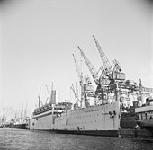 Photographie en noir et blanc d'un navire amarré à un quai, à côté de grues métalliques.