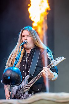 Kytarista Tommy Johansson během vystoupení se Sabaton v roce 2019. Stojí před mikrofonem, zpívá do něho a hraje na kytaru.