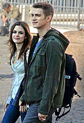 A woman and man, both smiling, are walking towards the left side of the image. The woman is wearing a white, button-up shirt and blue jeans. The man is wearing a green jacket, a blue shirt, a backpack, and blue jeans. In the background a man can be seen sitting and looking down.