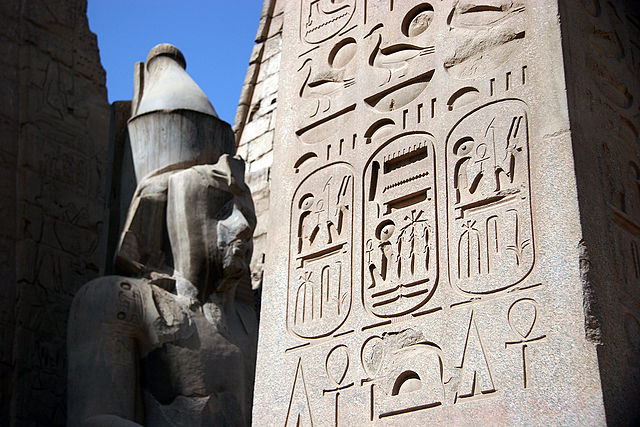 Incisioni geroglifiche che coprono un obelisco in primo piano. È presente una statua di roccia sul fondo.