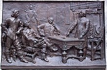 Photographie d'un bas-relief en bronze présentant plusieurs personnages autour d'une table, deux hommes se tenant debout, les deux autres assis.