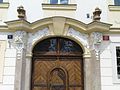Barokní portál s maskarony ve volutových pseudokonzolách po stranách a s nepříliš vydařenými rokokovými vázami (Hradčanské náměstí v Praze)