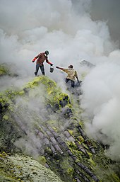 Traditional sulfur mining at Ijen Volcano, East Java, Indonesia. This image shows the dangerous and rugged conditions the miners face, including toxic smoke and high drops, as well as their lack of protective equipment. The pipes over which they are standing are for condensing sulfur vapors. Bergelut dengan asap nan beracun.jpg
