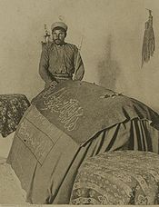 Фотография в оттенках сепии, на которой изображен мужчина в тюрбане, его левая рука держит тонкий посох, а он стоит позади и положил правую руку на кентотаф, задрапированный тканью, на котором есть надпись арабским шрифтом.
