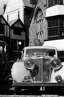 A black and white image of the front of an antique car, behind it can be seen portions of several old buildings