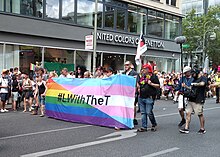 Photographie d'une manifestation où un groupe tient une banderole "L with the T" sur fond du drapeau arc-en-ciel et du drapeau trans