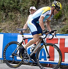 En cykelrytter i en blå og hvid trøje med gult finish uden nogen skrift på. Han står op i pedalerne op af et bjerg, mens en tilskuer betragter ham bag en barrikade.