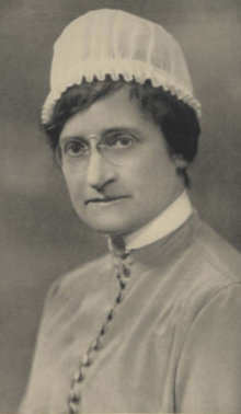 A middle-aged white woman wearing a nurse's uniform and cap, and glasses