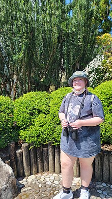 A picture of a person standing in front of various shrubs and bushes. They are smiling at the camera. Their hair is pink and they are holding binoculars.