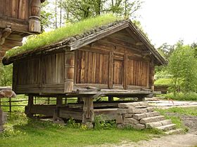 Una cabaña de maderas y leños al aire libre con un techo inclinado sobre un cimiento en piedras, con el segundo nivel volando del primero