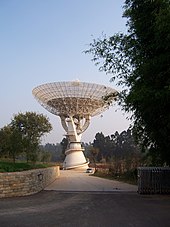 Photographie d'une superstructure au milieu d'un paysage arboré, formée d'un support élancé et d'une couronne parabolique tournée vers le ciel.