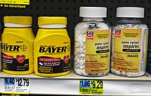 Четыре пластиковых бутылки с лекарствами на другой полке аптеки над ценниками. Два слева желтого цвета со словом «Байер», выделенным черным шрифтом; Выше небольшой шрифт описывает продукт как «настоящий аспирин». Слева видны две прозрачные пластиковые бутылки с логотипом сети аптек Rite Aid на желтых этикетках, которые описывают продукт как «обезболивающий аспирин».