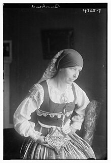 A white woman seated indoors at a window, wearing a folk costume with a headscarf