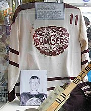 White hockey jersey with maroon trim and a Bombers logo on the chest, accompanied by a hockey stick, a hockey glove, and a photo of Bobby Clarke