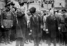 Paul Deschanel salue la foule de son haut-de-forme ; derrière lui, les officiels se tiennent en rangs serrés.