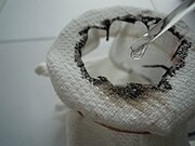 Biała bawełniana tkanina, na którą pipetą nanoszony jest kwas siarkowy; widoczna jest dziura ze zwęglonymi brzegami.