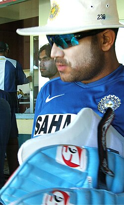 A man in the blue Indian cricket practice kit,wearing sun cream,sunglasses and a hat carrying his batting pads. Others can be seen in the backgrounf.