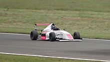 Photographie d'une monoplace de Formule 4 blanche et rouge, vue de trois-quarts droit, sur une piste sèche, entourée par des bandes d'herbe.