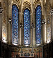 Photographie en couleurs de la chapelle vue de face avec au centre trois vitraux, de part et d'autre une mosaïque murale et en bas le retable.