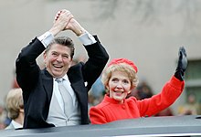 À travers le toit d'un véhicule noir, un homme en costume souriant se tient les mains au-dessus de la tête dans un signe de victoire. À ses côtés, une femme habillée de rouge salue de la main gauche.