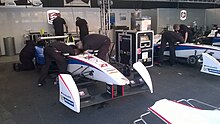 Photo d'un stand de course, où deux mécaniciens travaillent sur le cockpit d'une monoplace