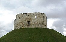 A photograph of a small castle on top of a green mound; the castle has three circular walls visible. Behind the castle the sky is overcast and dark grey.